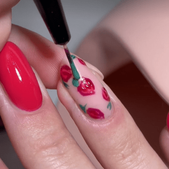 A close-up of a hand painting a nail with a brush, with the nail decorated with red flowers and green leaves on a pink background.