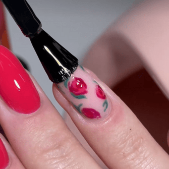 A nail with rose nail art is covered with a bright top coat by a brush, next to a bright red painted nail.