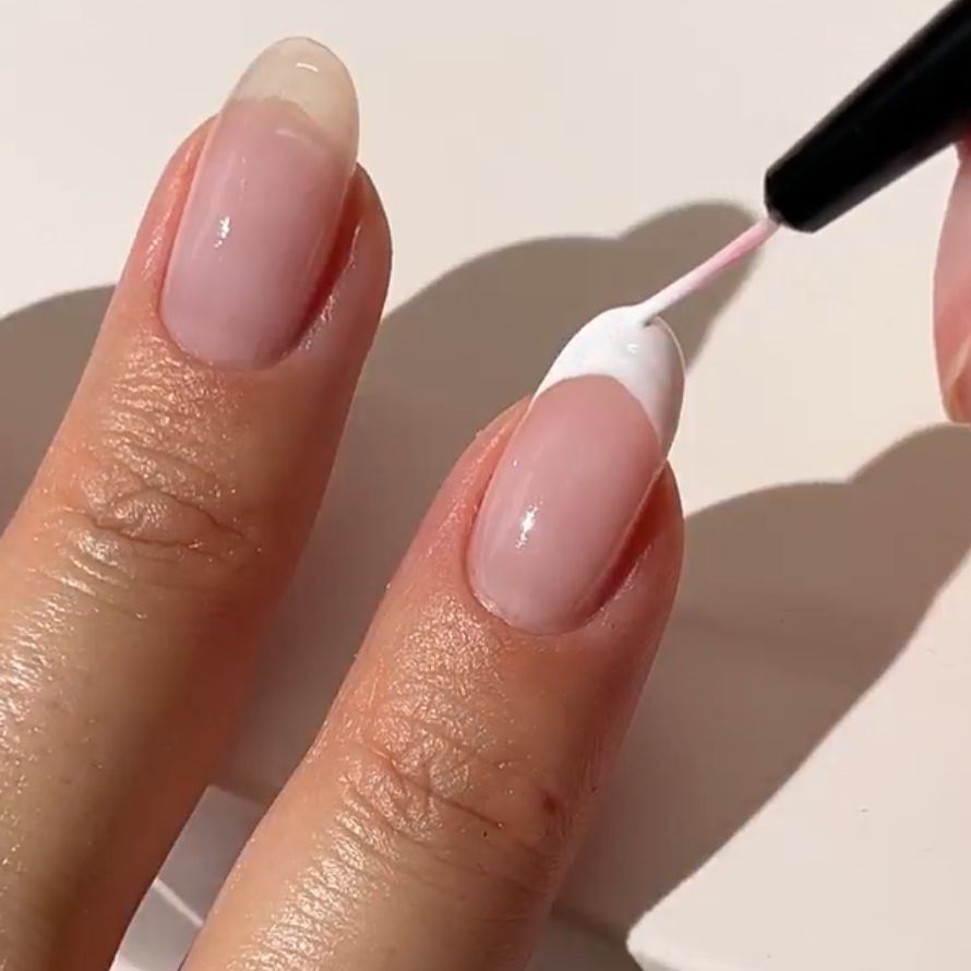 A hand where the nails are painted with a white nail polish, focus on the fingers and the brush.