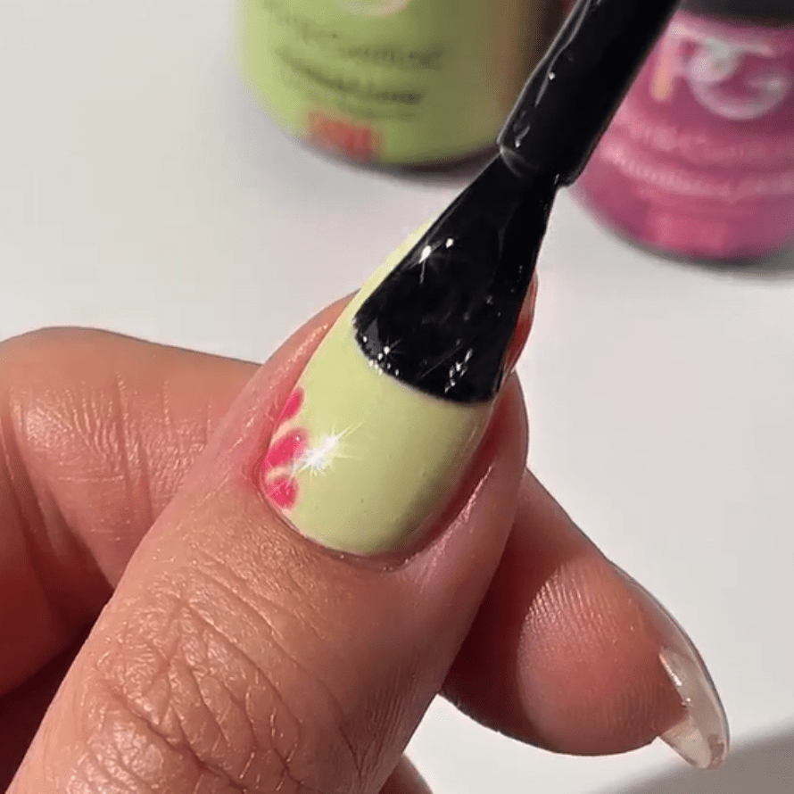 A hand showing a nail with a light green polish and a pink floral design, while a brush goes over it for a smooth finish. In the background are some colored nail polish bottles.