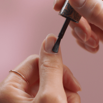 A close-up of someone applying nail polish to a thumbnail against a pink background.