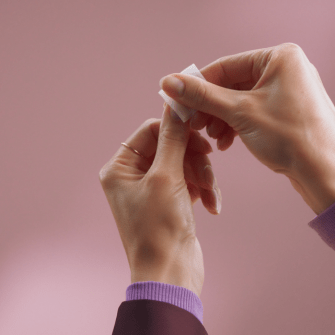 Two hands against a pink background, with one of the hands holding a small square cloth.