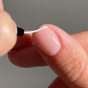 A close-up of a hand applying a subtle white line to a natural nail using a thin brush.