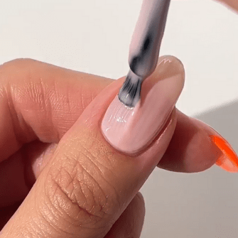 A hand paints a nail with a light pink nail polish on a neutral background.