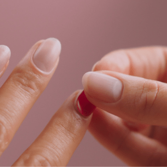 A person holding a red capsule on a finger against a pink background.