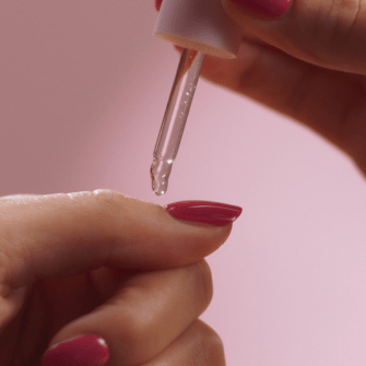 A person applies a drop of oil to a nail using a pipette, with a pink background.
