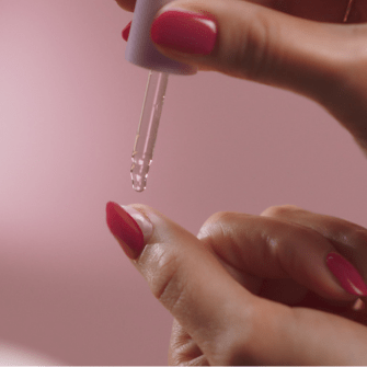 One hand applies a drop of liquid with a pipette to another hand with pink painted nails, against a pink background.