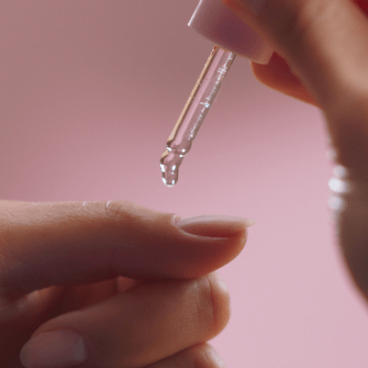 Two hands hold a pipette from which a drop of liquid falls onto a fingernail, against a pink background.