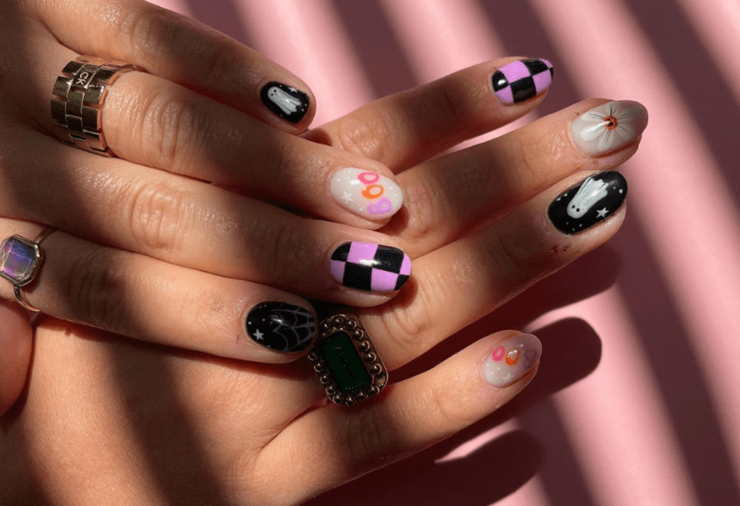 Hand with creative nail art: ghosts, checkerboard pattern, spider web and dots on shiny nails, worn with rings.