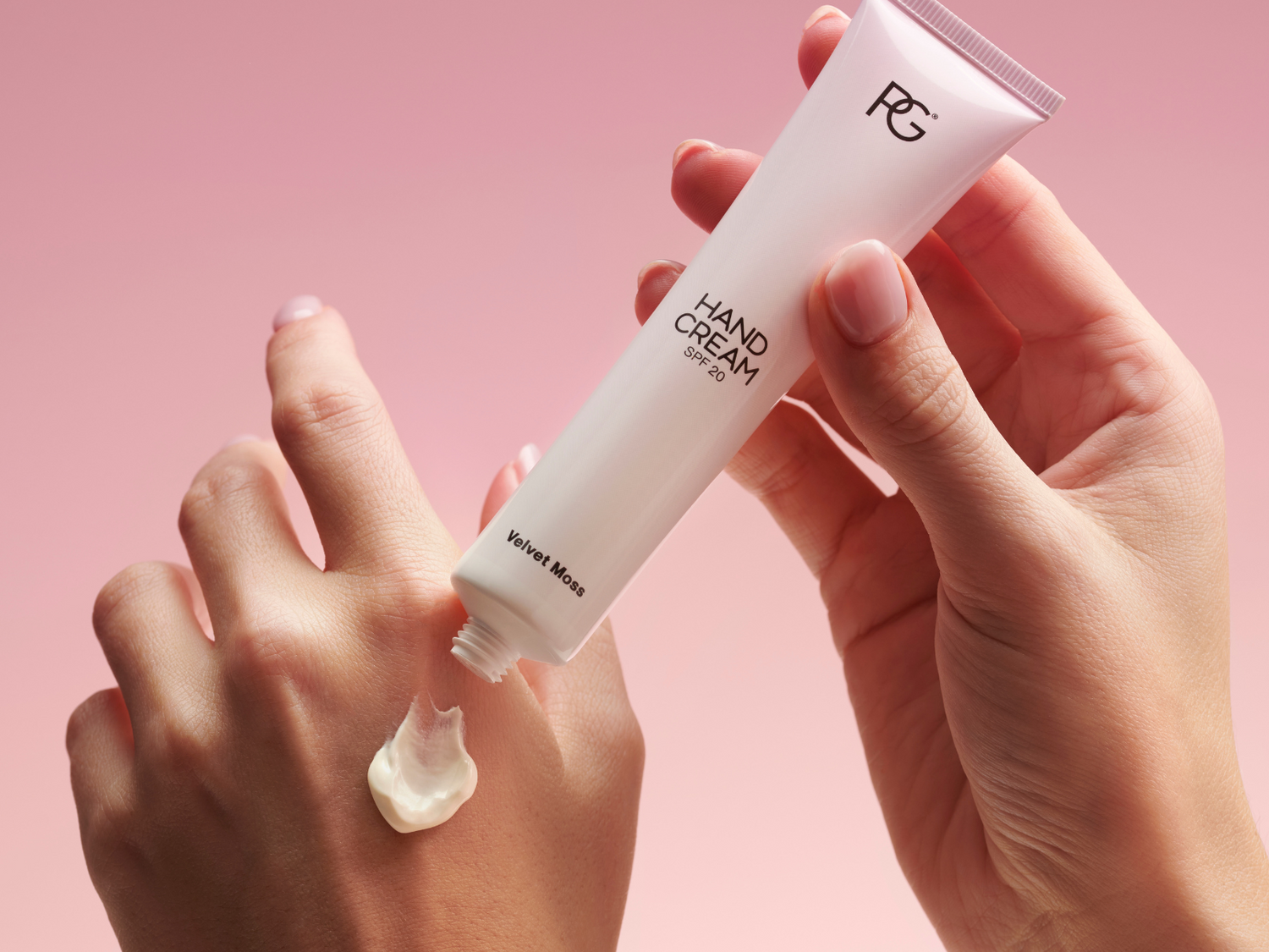 Two hands, one©©squeezes a tube of hand cream above the other hand, on a pink background.