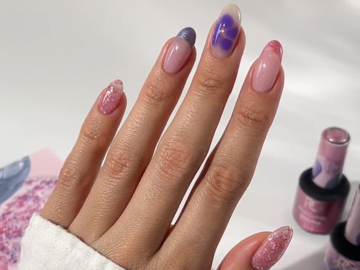 Hand with nails in pink and purple nail art designs, with glitter and marble effects. In the background bottles of nail polish.