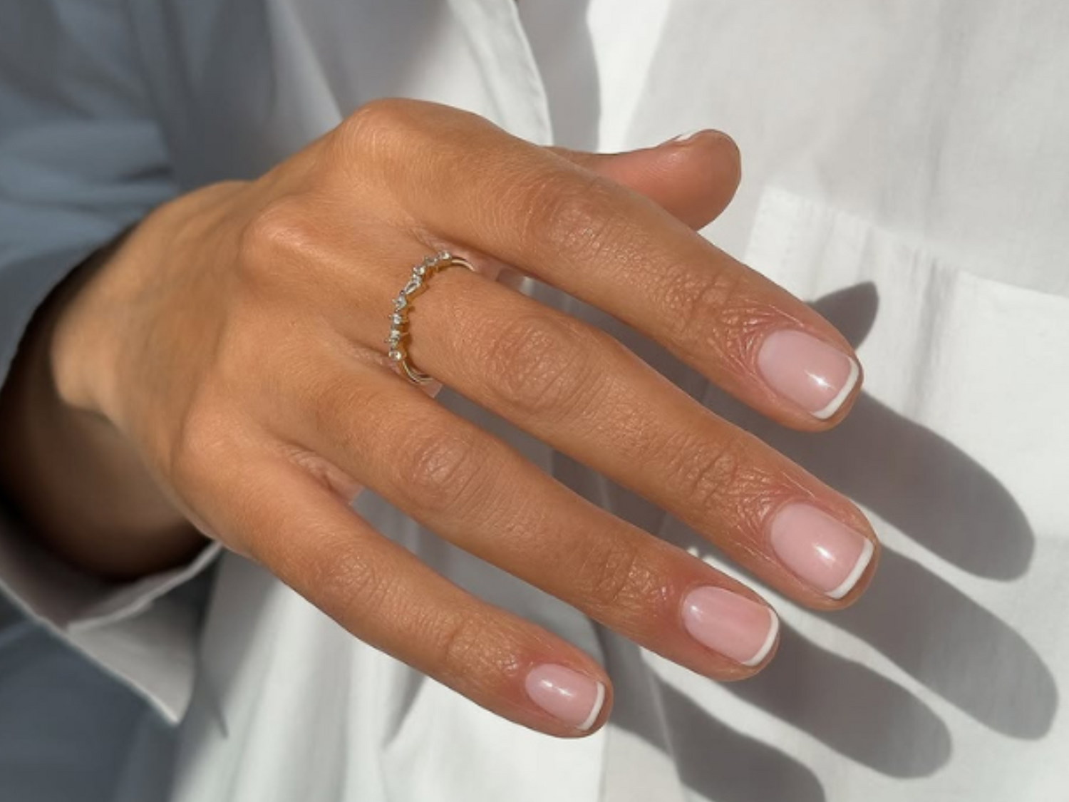 A hand with a gold-colored ring set with small stones, with well-groomed nails in a French manicure style, against a white background.
