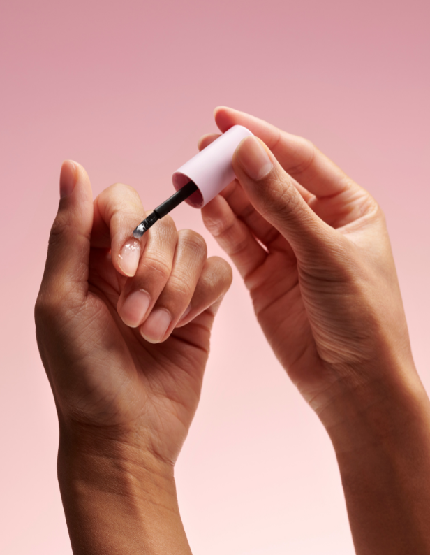 Two hands against a pink background where one hand paints the nail of the other hand with a light pink nail polish.