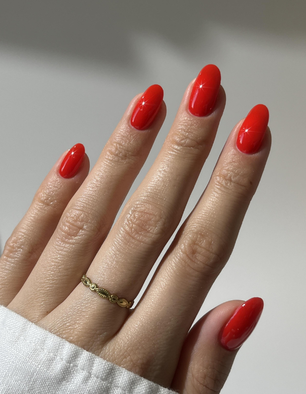 A hand with long, shiny, bright red nails and a gold ring on the ring finger against a neutral background.