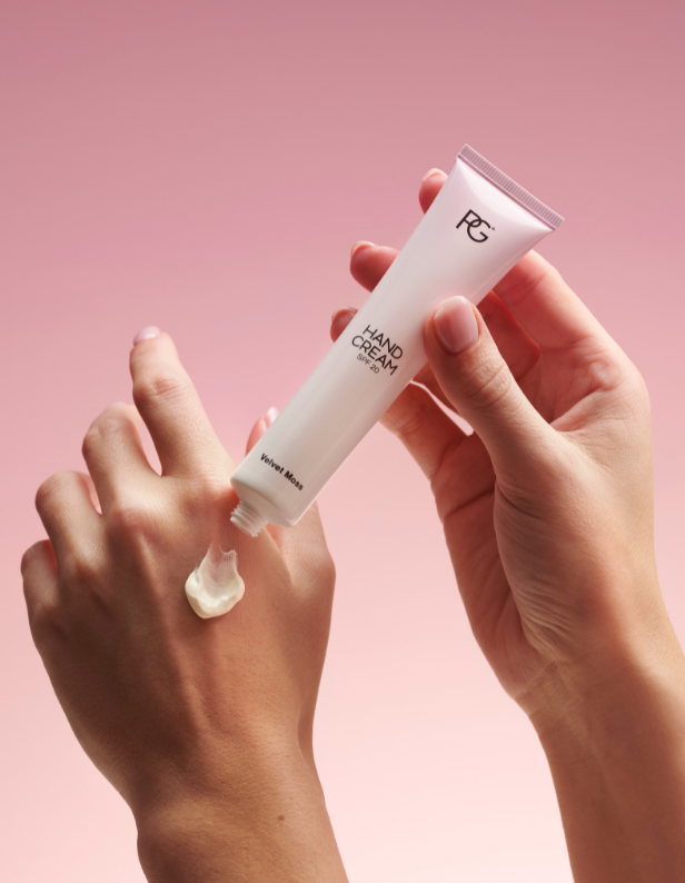 Hands apply a white hand cream to the back of a hand against a pink background.