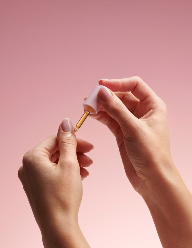 Hands applying a dropper with oil to a thumb against a pink background.