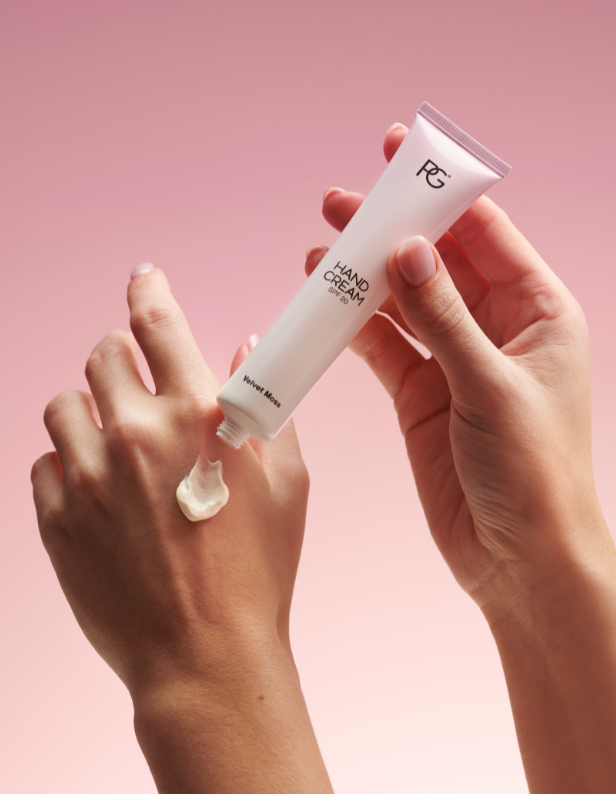 A person squeezes hand cream from a tube on the back of their hand against a pink background.