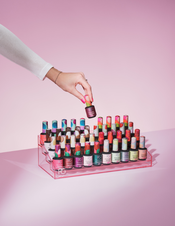 A hand reaches for a bottle of nail polish from a series of colorful nail polish bottles arranged on a transparent holder, against a pink background.