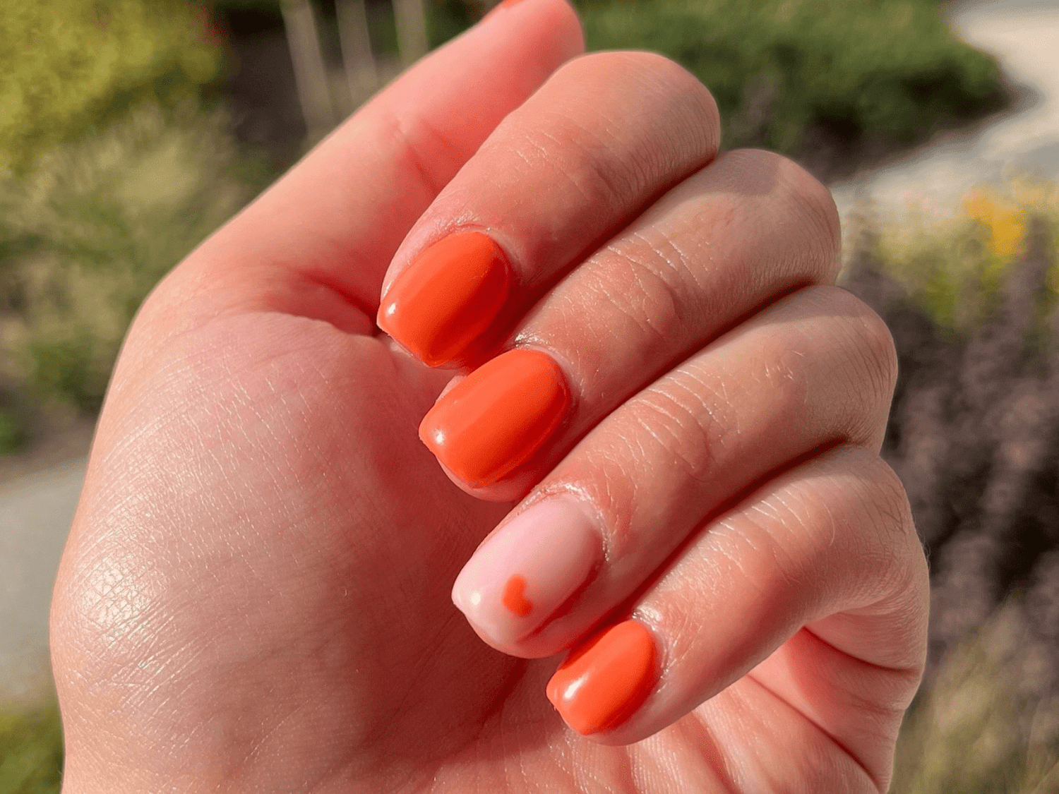 Close-up of a hand with bright orange nail polish on four nails and a light pink nail with a small orange heart.