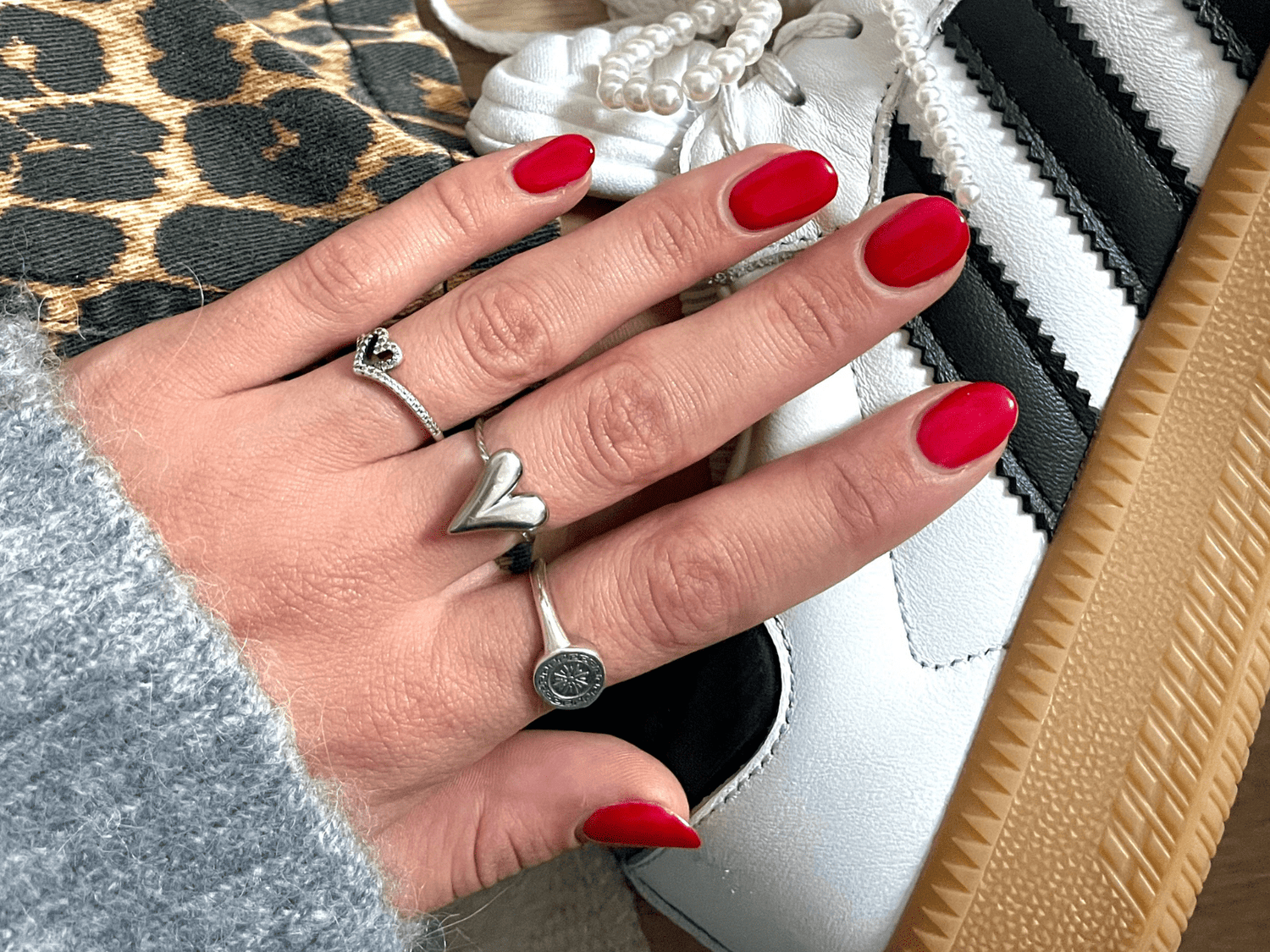 A hand with bright red nail polish wears rings in the shape of hearts, resting on a white sneaker with black stripes and brown sole, with a background of leopard print fabric.