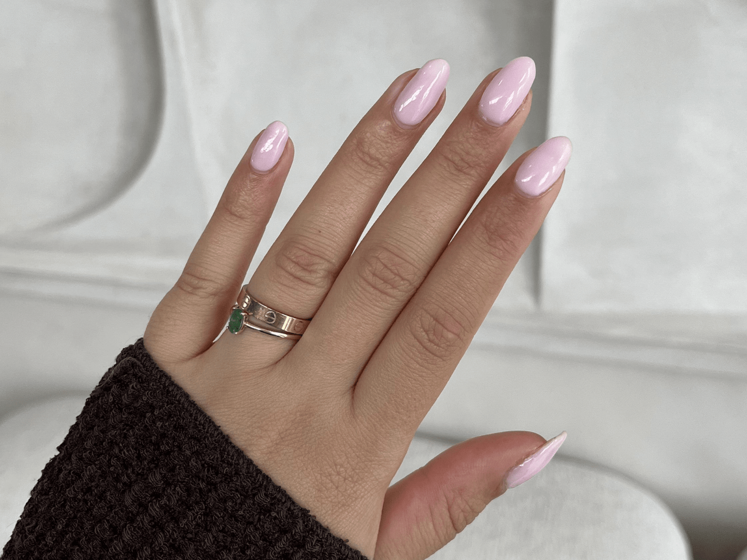 A hand with pink painted nails, two rings on the ring finger, one©©of which has a green stone, against a neutral background.