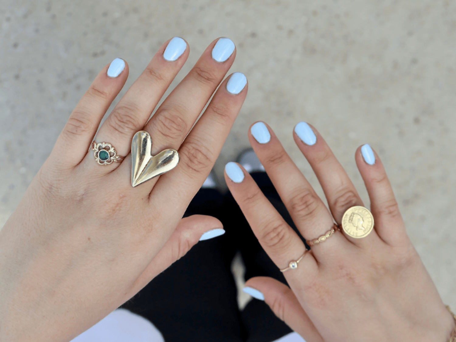 Two hands with light blue painted nails, decorated with several rings, including a heart-shaped one and one with a coin design.