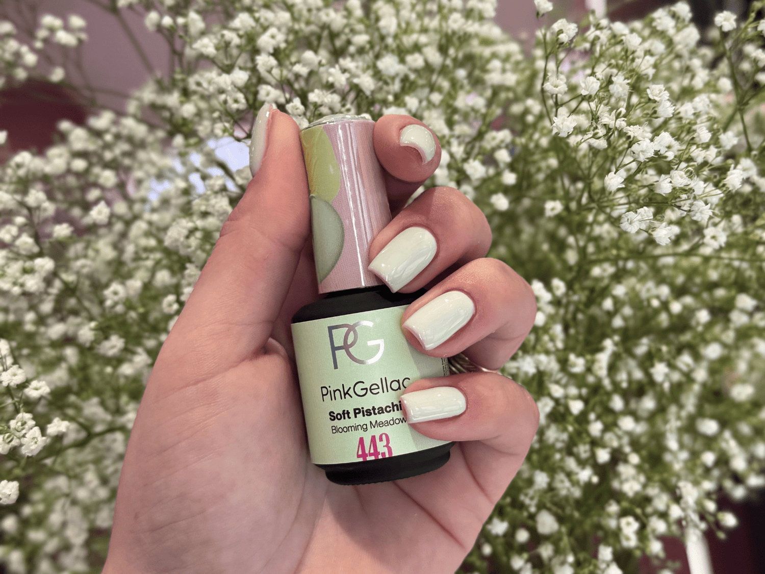A hand with painted nails in a soft pistachio color holds a bottle of Pink Gellac nail polish, against a background of white flowers.
