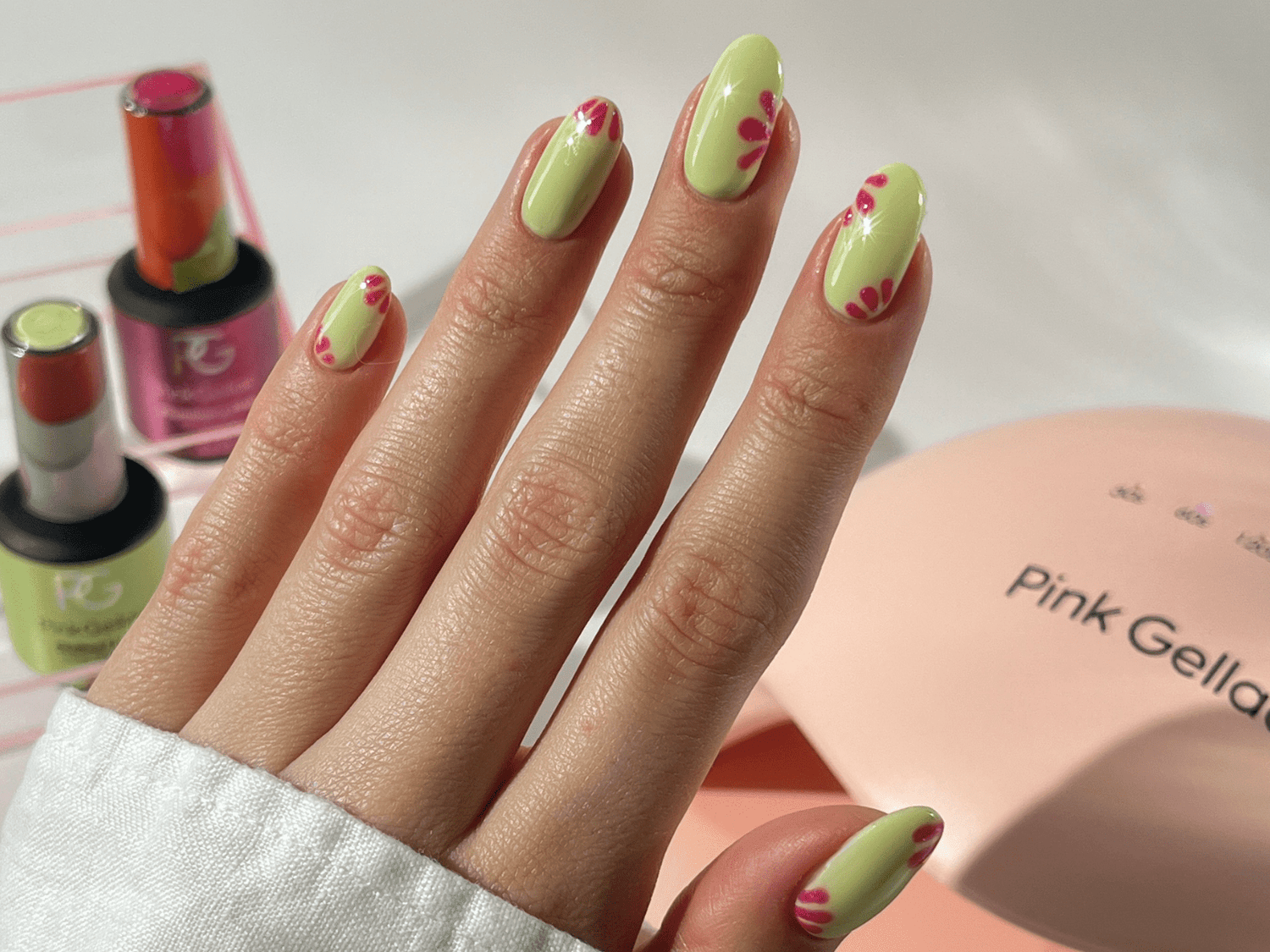 A hand with light green nail polish and pink floral design, next to some bottles of nail polish and a pink nail dryer with the text "Pink Gellac".
