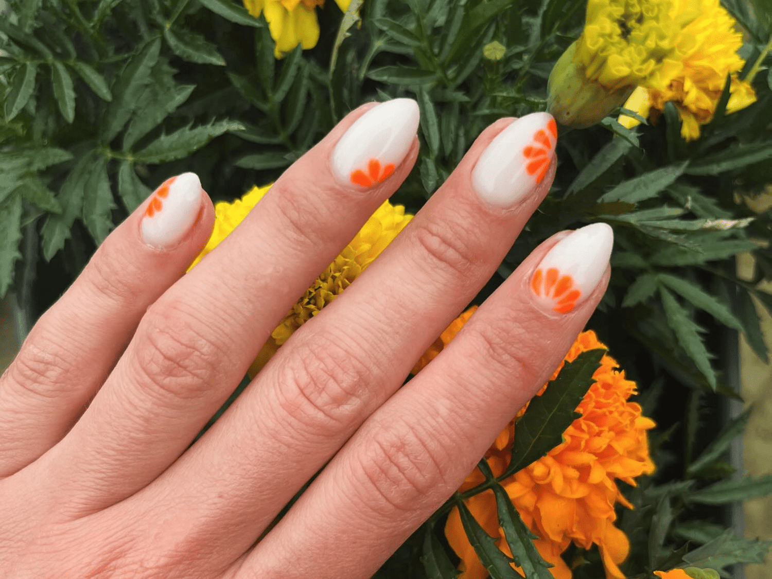 A hand with a manicure of white nails with orange floral motifs, against a background of orange and yellow flowers.
