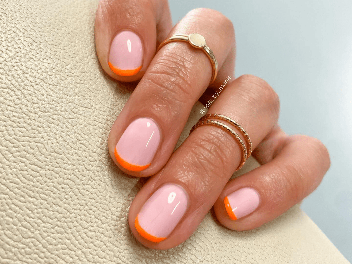 A hand with pale pink nails with an orange French manicure tip, three gold rings and a cream structure background.