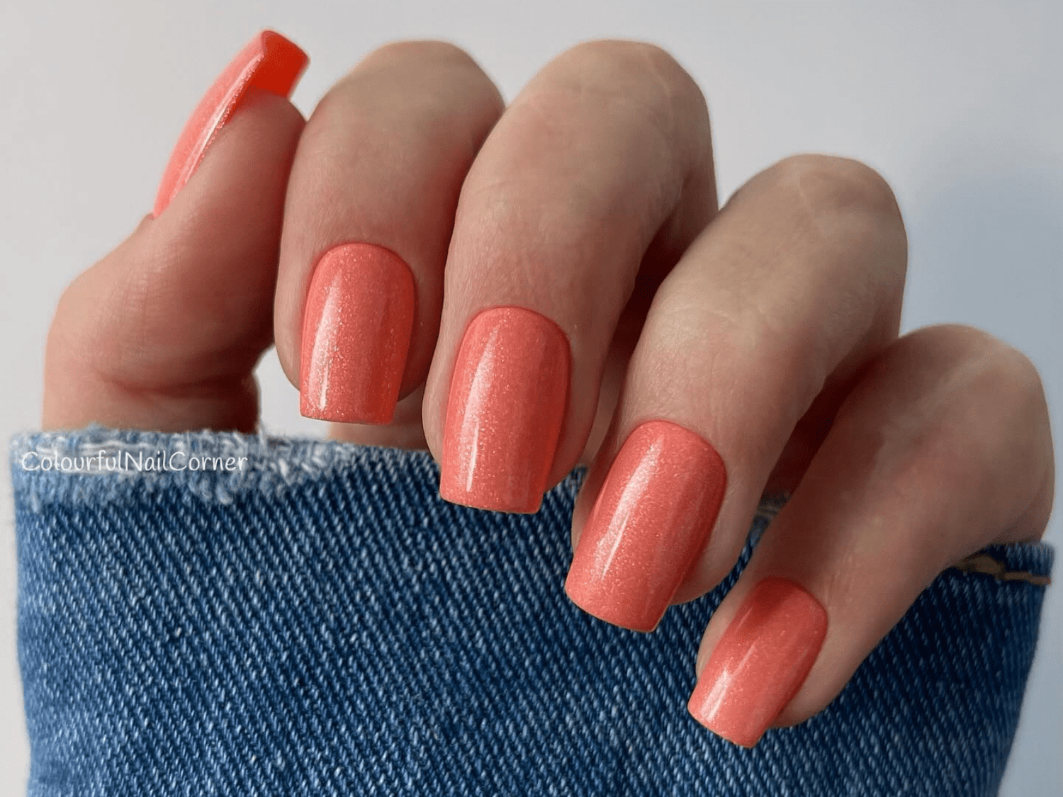 Hand with long nails painted in coral color, partially covered by a denim sleeve.