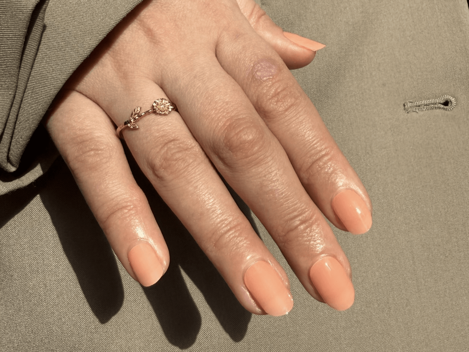 A hand with peach nail polish and a detailed ring with a flower and leaf design on an olive green fabric.
