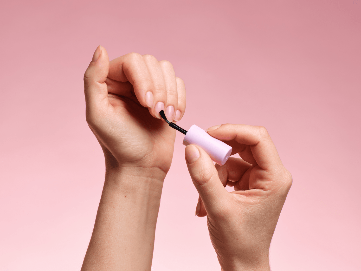 Hand paints nails with clear polish in front of pink background.