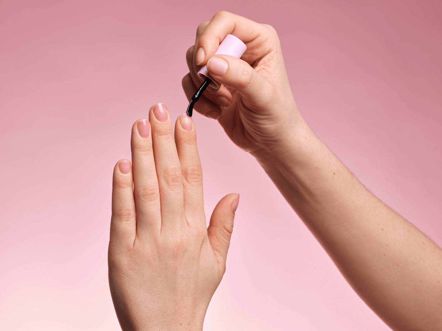 Hand paints nails with black nail polish against a pink background.