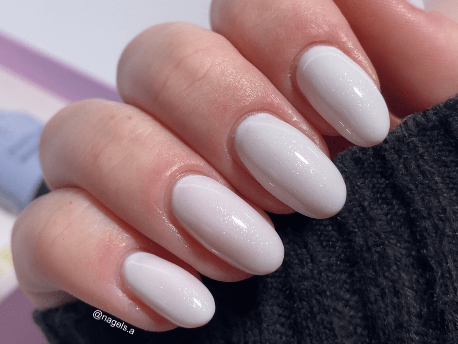 White, shiny artificial nails on a hand, worn with a dark sleeve in the background.