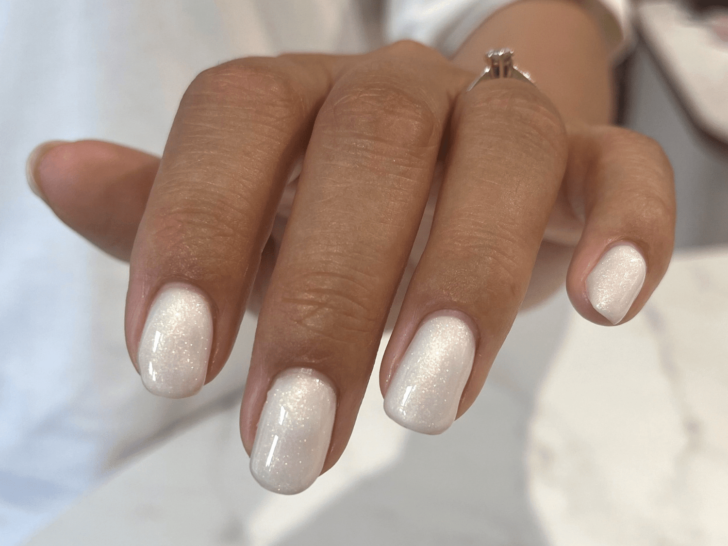 A hand with short, white shiny nails and a ring on the ring finger.