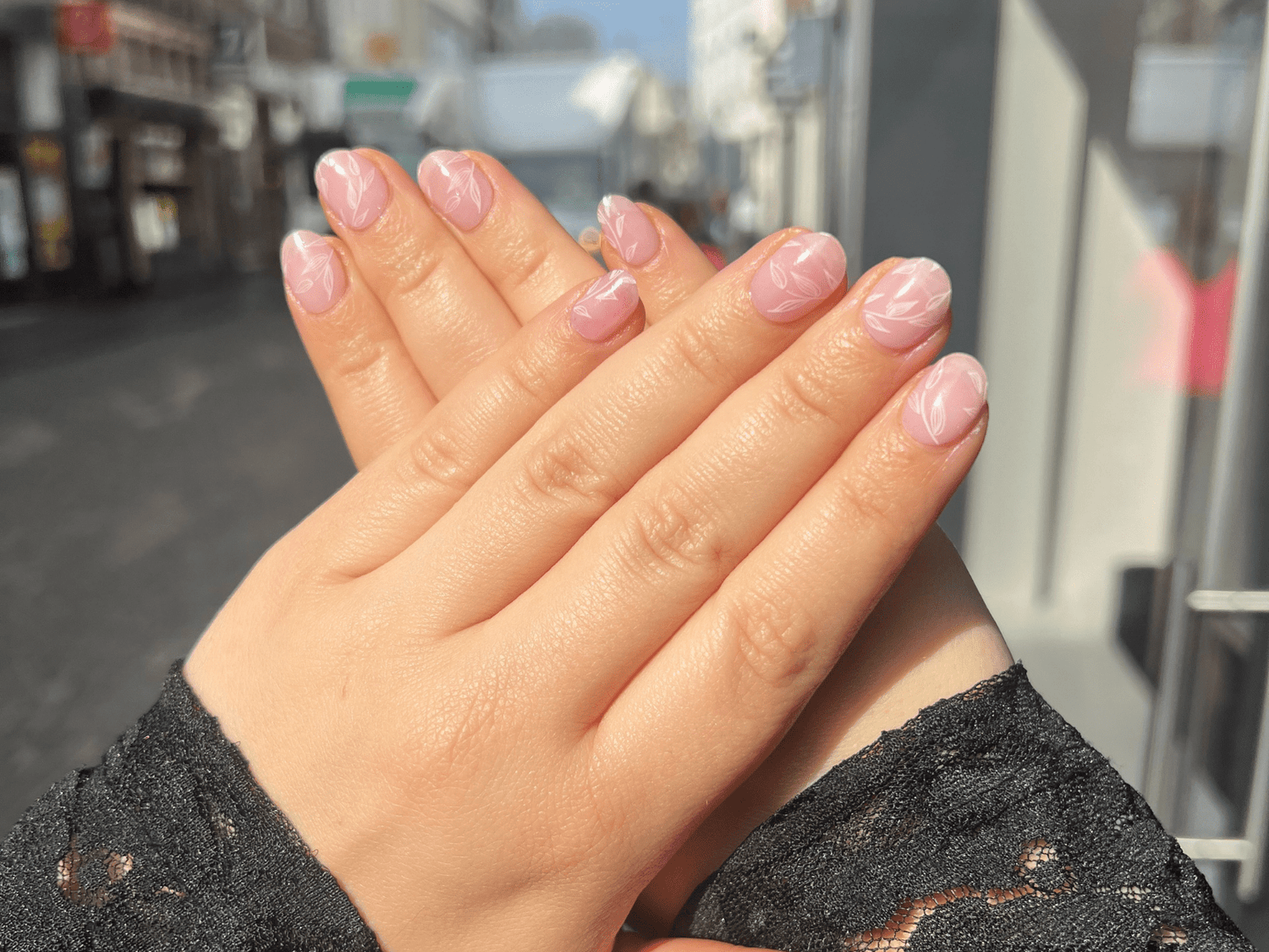 Hands with pink nail polish and white floral motifs, against a blurred urban background.