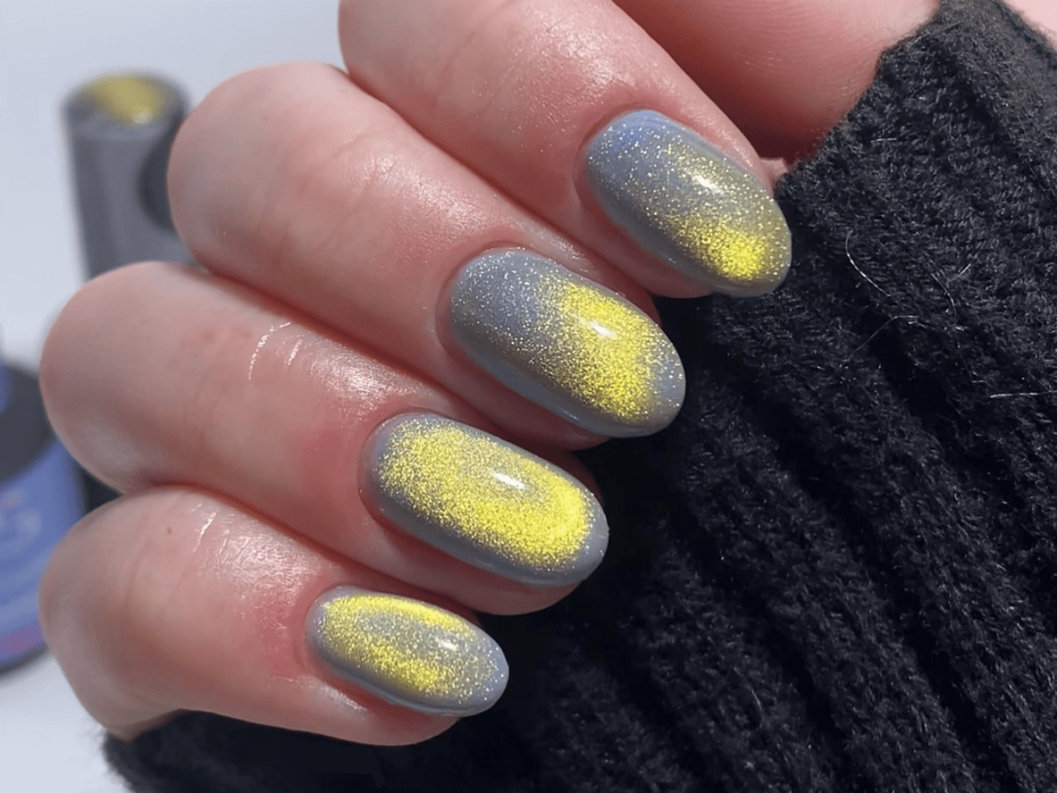 Gray with yellow nails, glitter effect, worn by a hand in a dark sweater.
