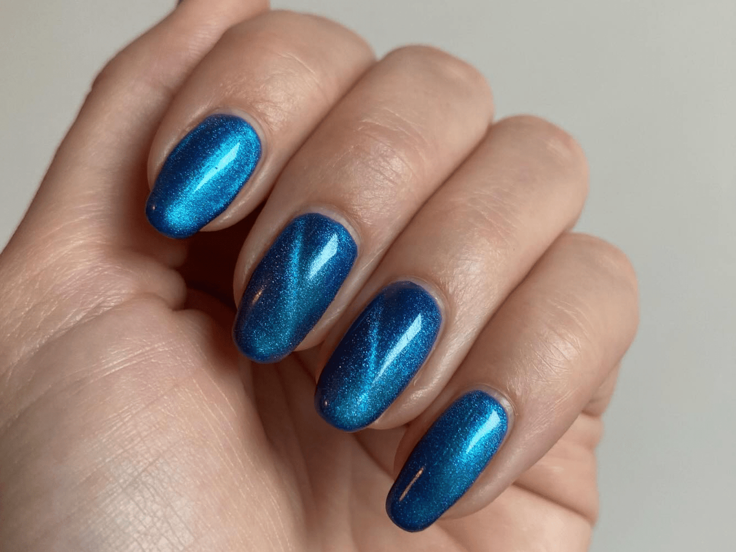 A hand with blue metallic painted nails, shiny and well-groomed against a neutral background.