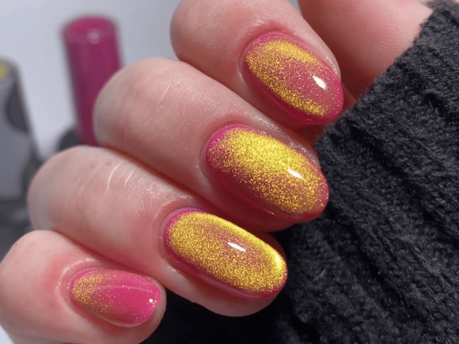 Nails with pink and yellow glitter gradient, worn by a hand with black sleeve.
