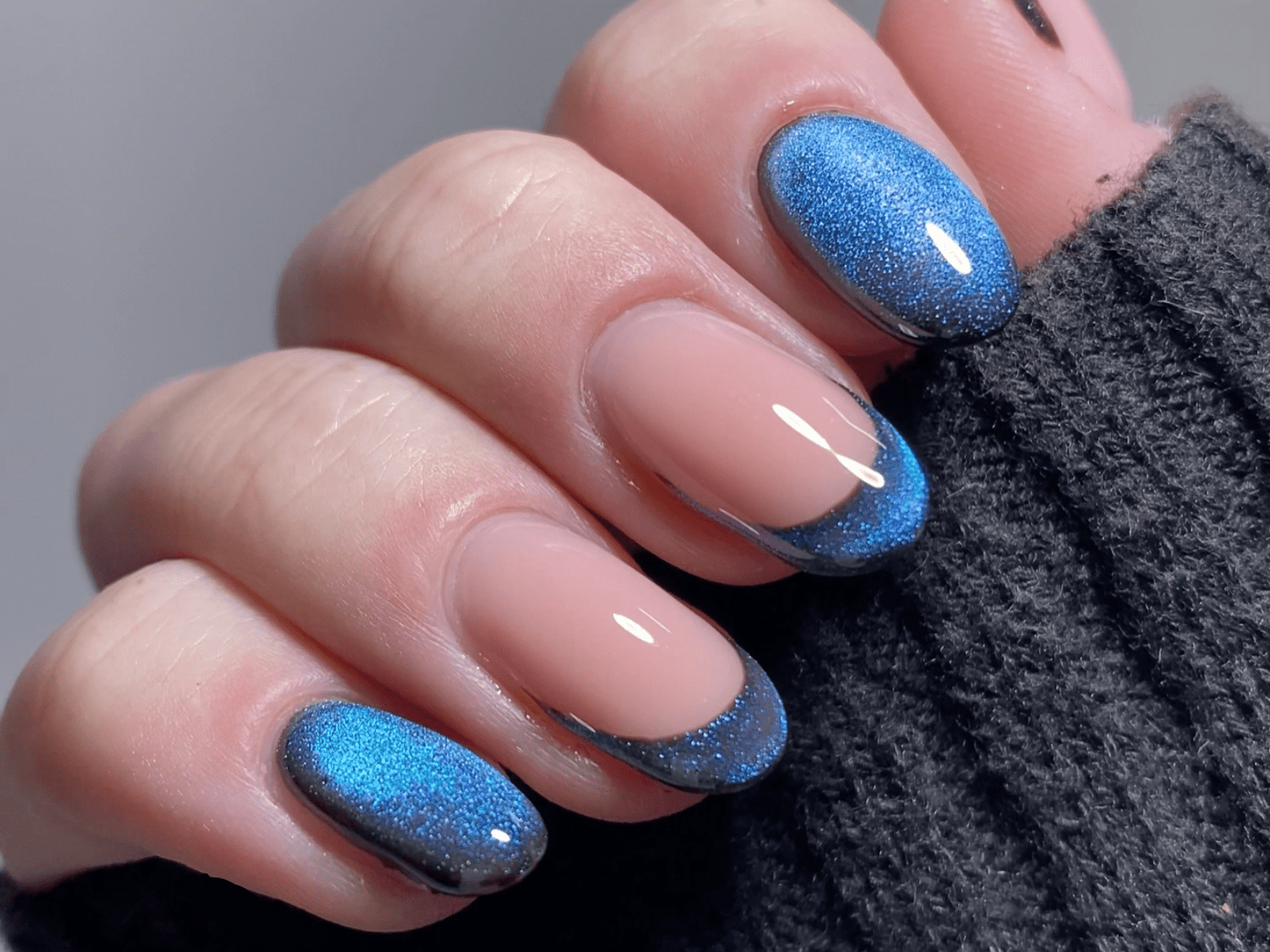 Hand with nails in shiny blue-black ombre style, worn by a person in a black sweater.