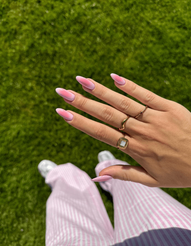 A hand with pink nails and several rings, facing the camera, with green grass in the background and under striped pink and white pants and white shoes.