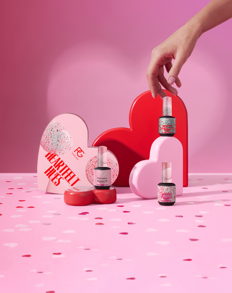 An image with pink and red heart-shaped boxes, nail polish bottles with glitter, and a hand holding a bottle on a pink background with confetti.