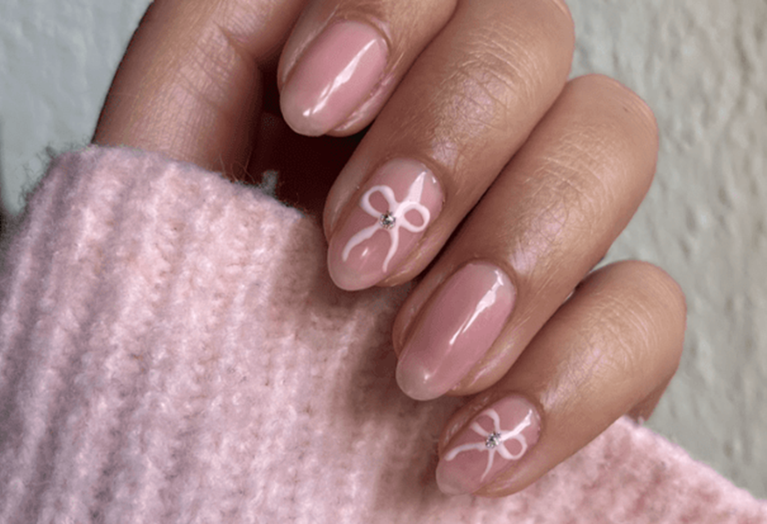 A hand with shiny, light pink nail polish, decorated with a subtle white bow and a stone on two nails, against a background of a pink knitted sweater.