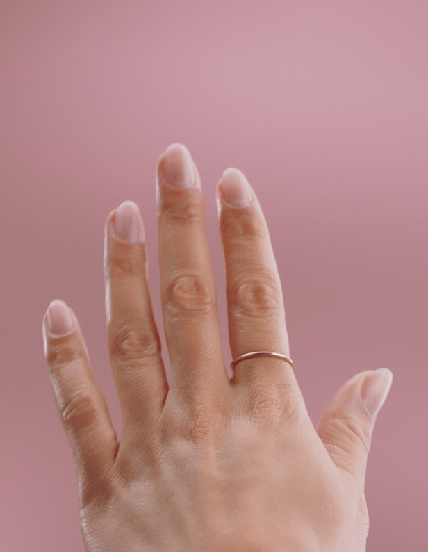 A hand with a simple gold ring on the ring finger against a pink background.