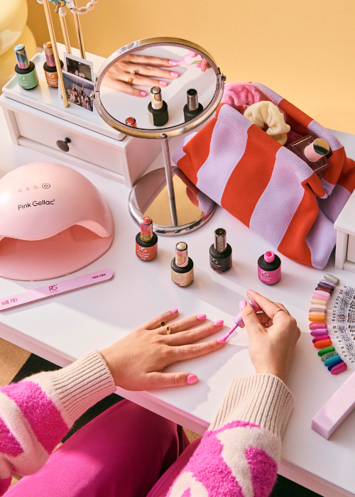 Person at a desk painting their nails pink with various nail polishes, UV lamp, mirror and accessories in the background.