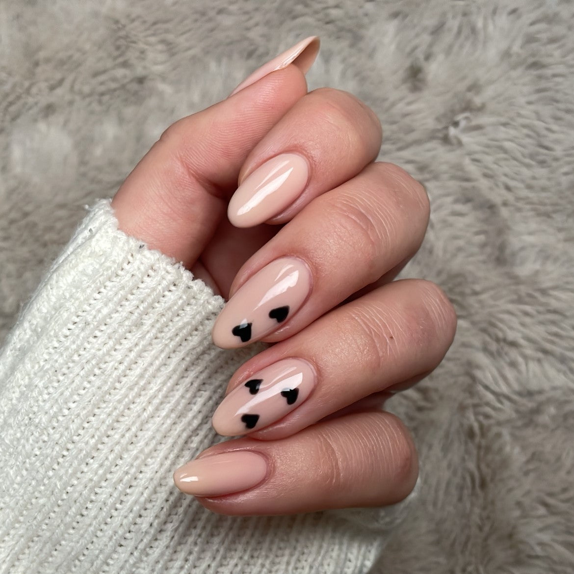 A hand with beige nail polish with black hearts on two nails, against a white sweater and gray background.
