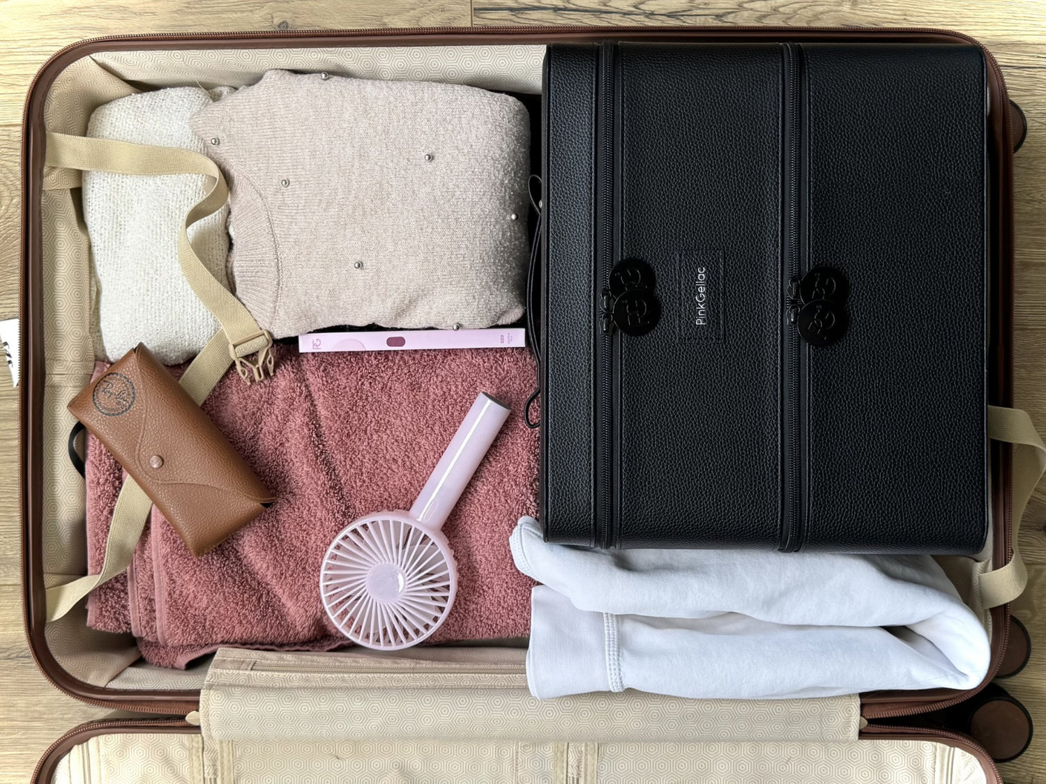 An opened suitcase containing clothes, a brown glasses case, a pink hand fan and a black makeup organizer.