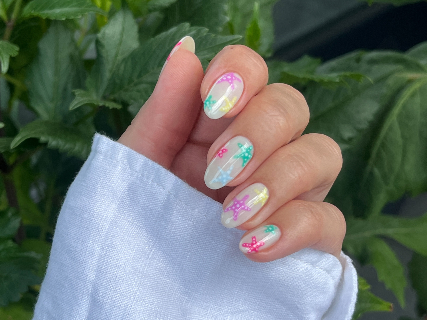Hand with colorful nails, decorated with star-shaped patterns against a background of green leaves.