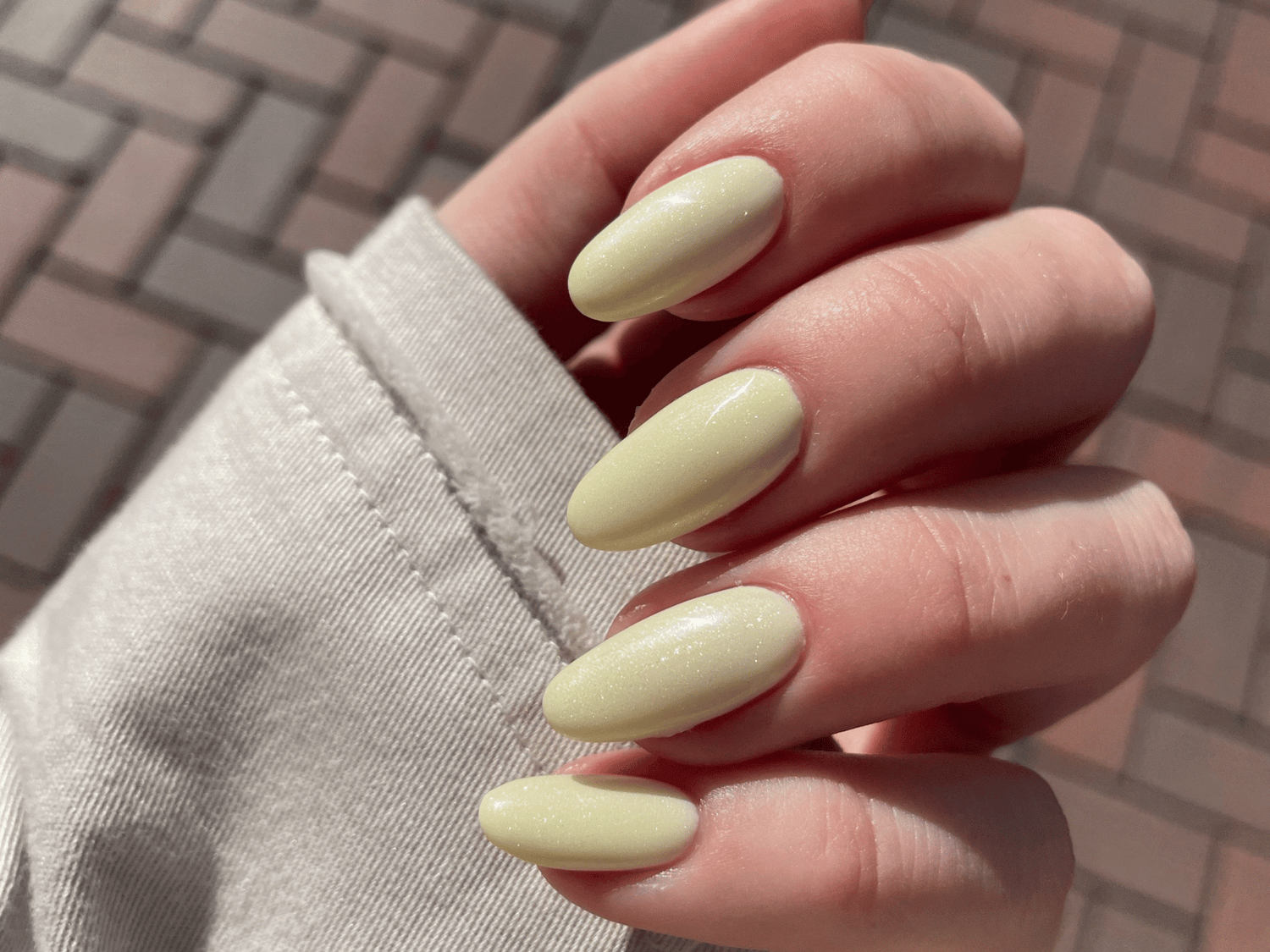 Hand with pastel yellow nail polish, glittering in sunlight, against a checkered stone background.